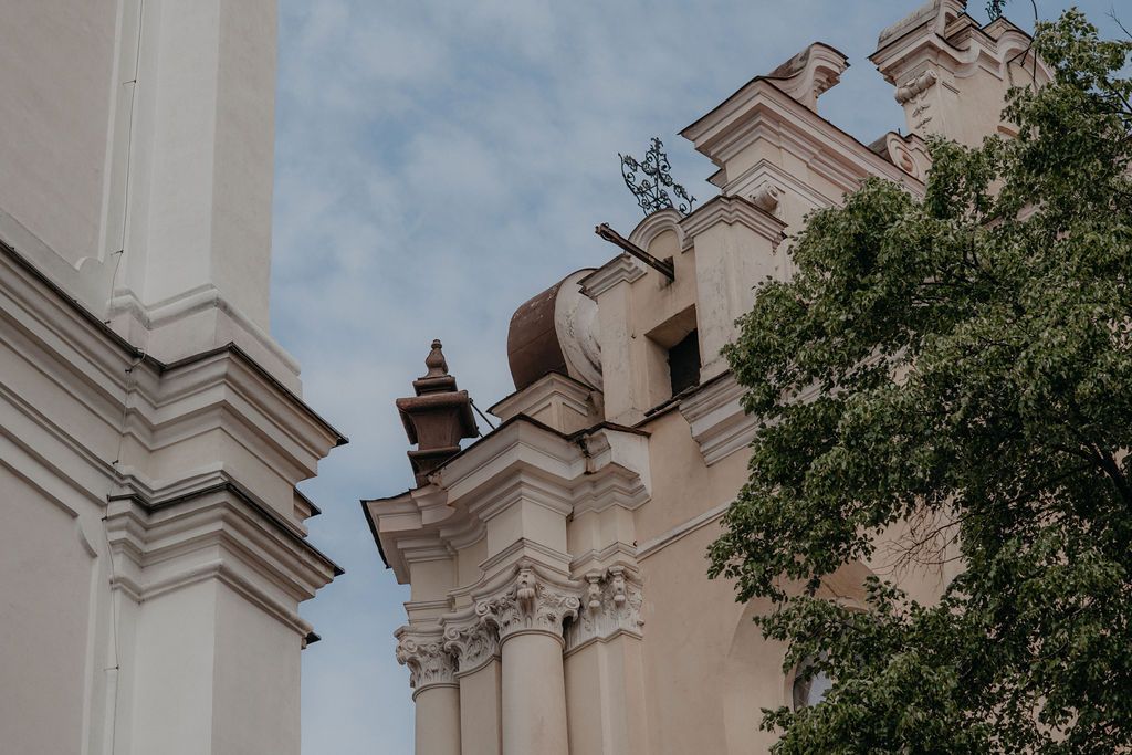 Historic baroque church facade with ornate white columns, decorative crosses, and lush green trees against a cloudy sky.