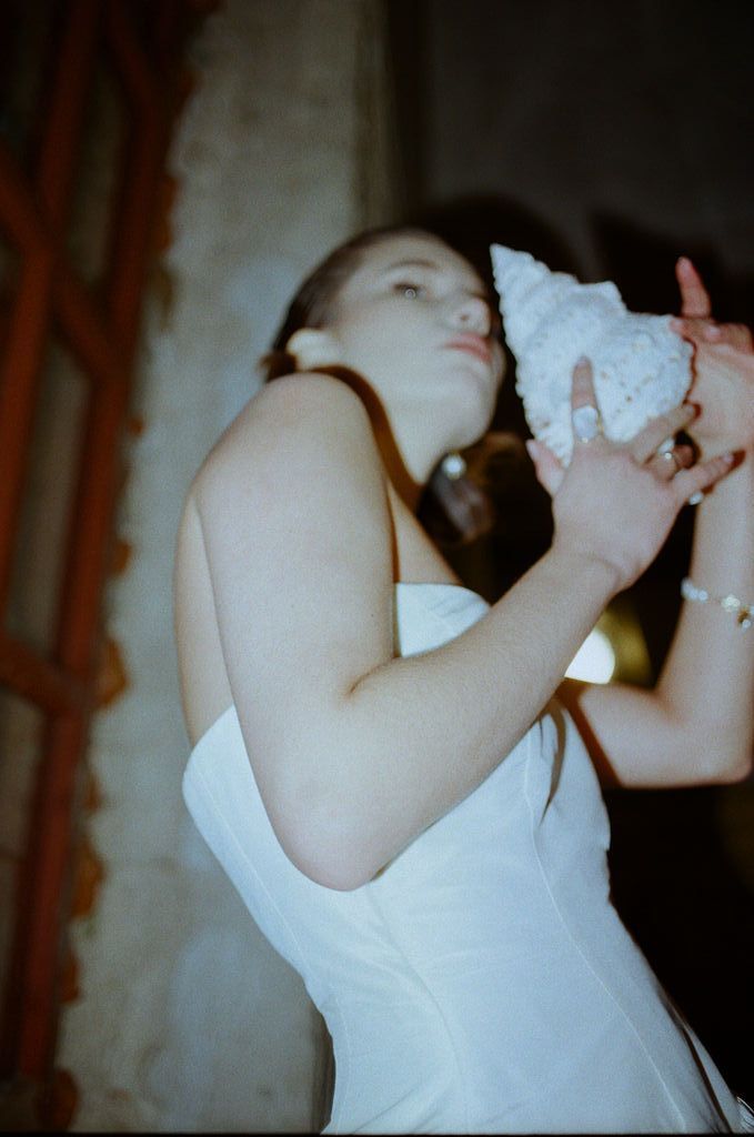 Bride in strapless white gown holding a large white conch shell to her ear in a softly lit venue.