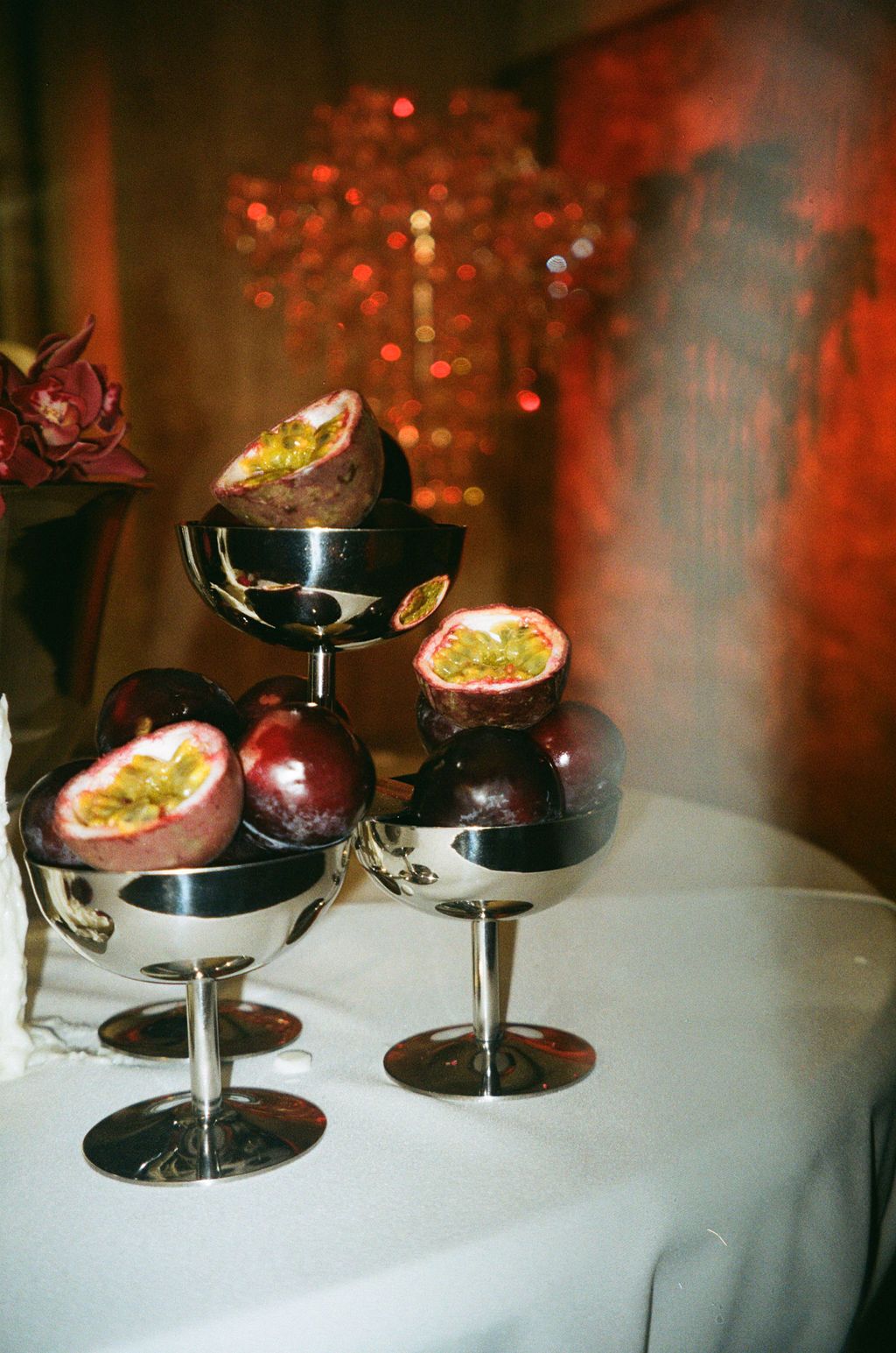 Silver pedestal bowls display fresh passion fruits, some halved to reveal seeds, arranged on a white tablecloth with red bokeh lights.