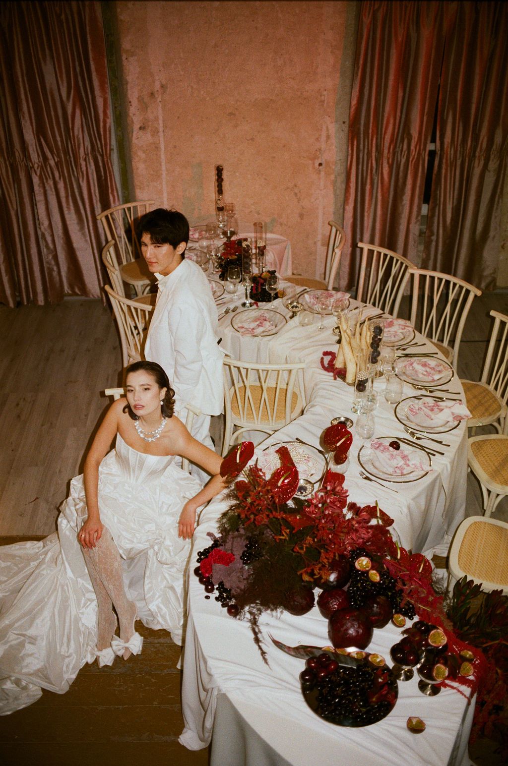 Bride in white gown and groom in white suit pose at elegantly decorated wedding table with red floral arrangements.