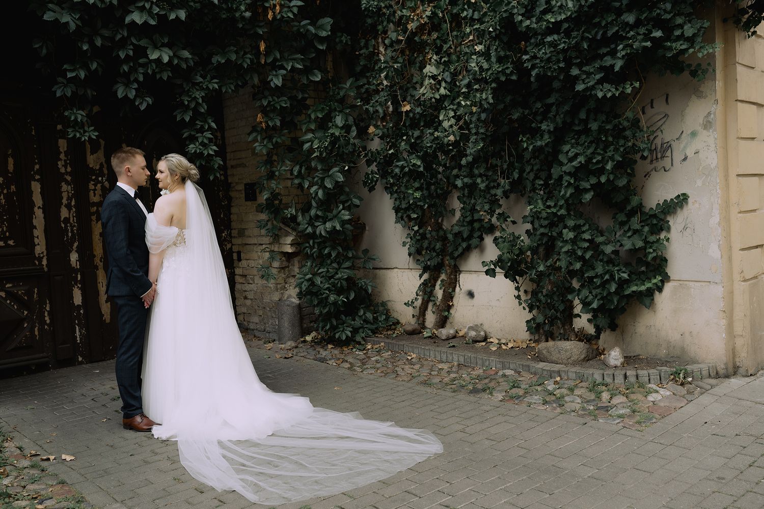 A couple in wedding attire shares an intimate moment in a lush garden setting with trailing white dress details.
