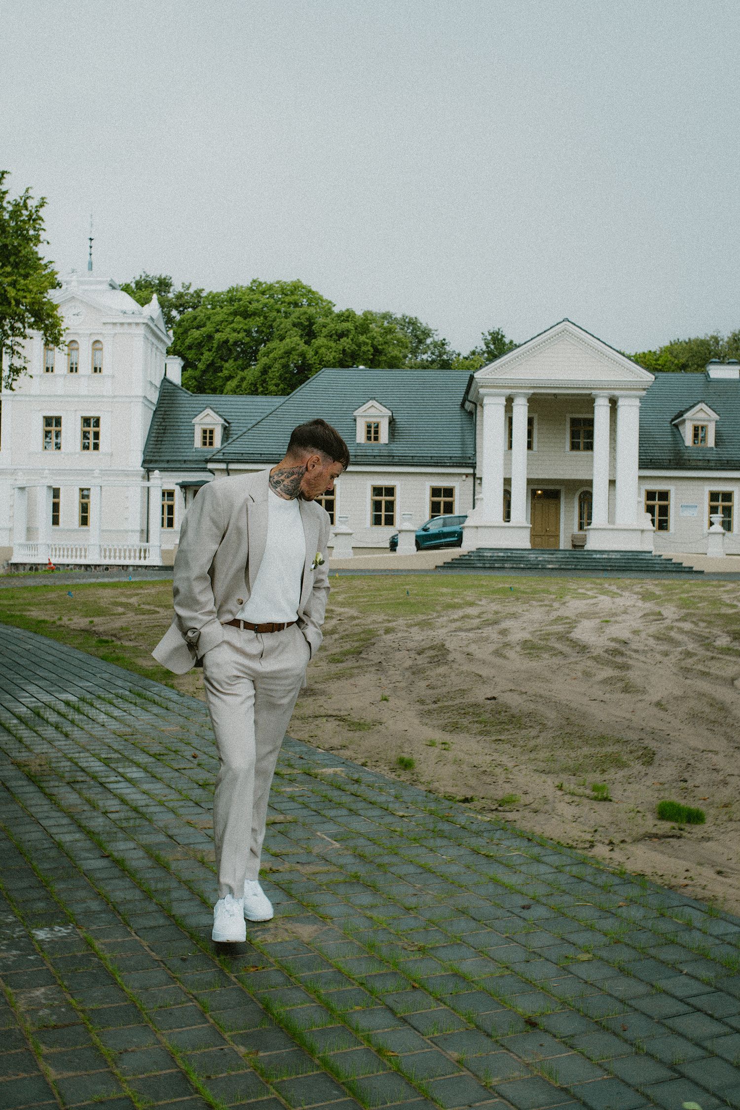 A person in a light gray suit walks across a paved courtyard with white colonial-style buildings in the background.