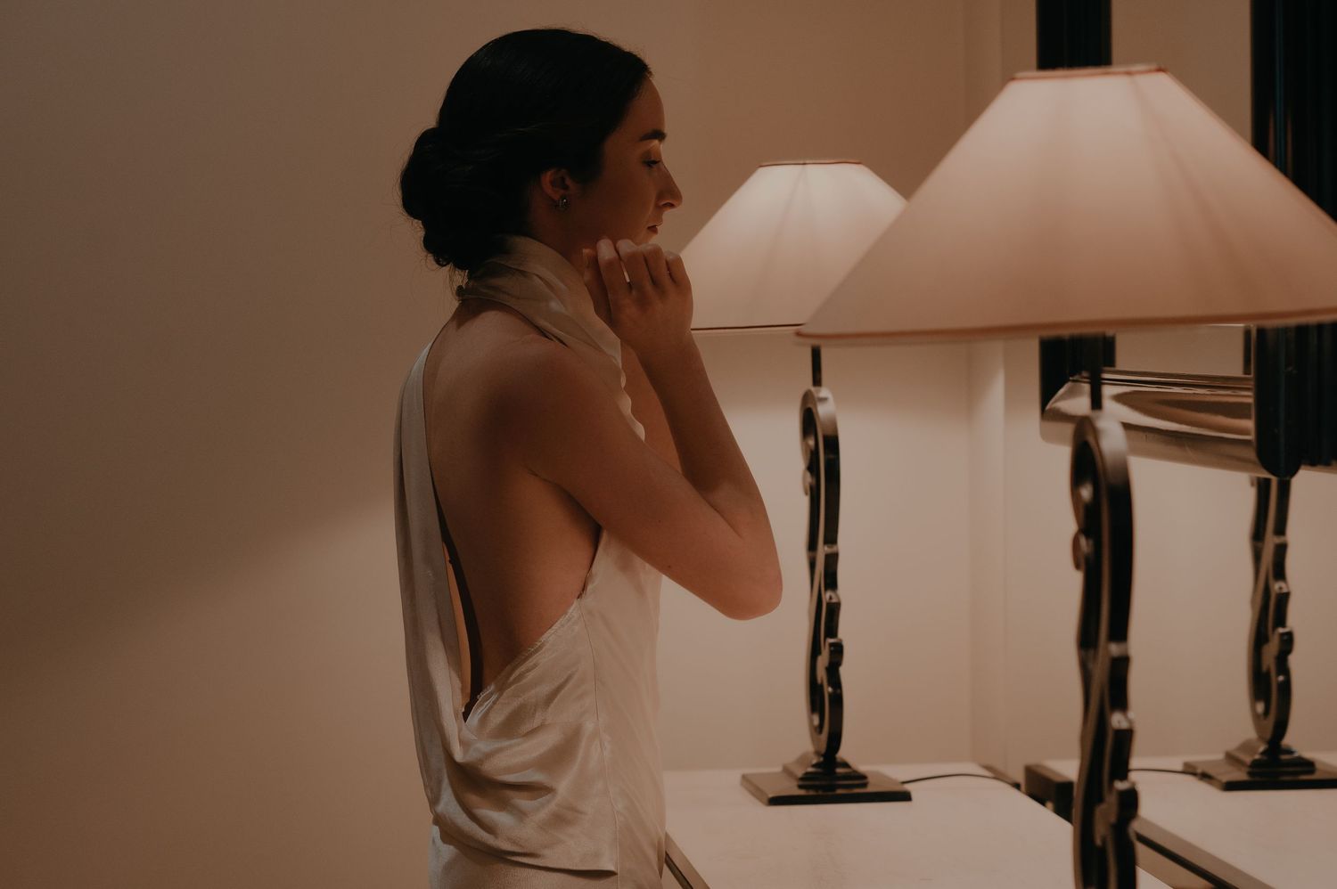 A person in a sleeveless top stands beside a table lamp in warm lighting.