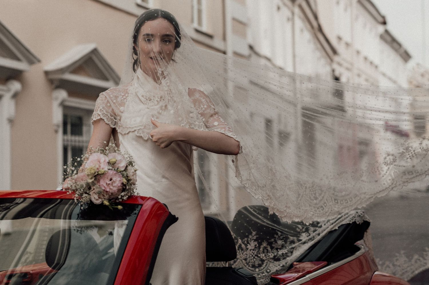 A bride in a flowing white dress sits in a classic red vintage car during a wedding photoshoot.
