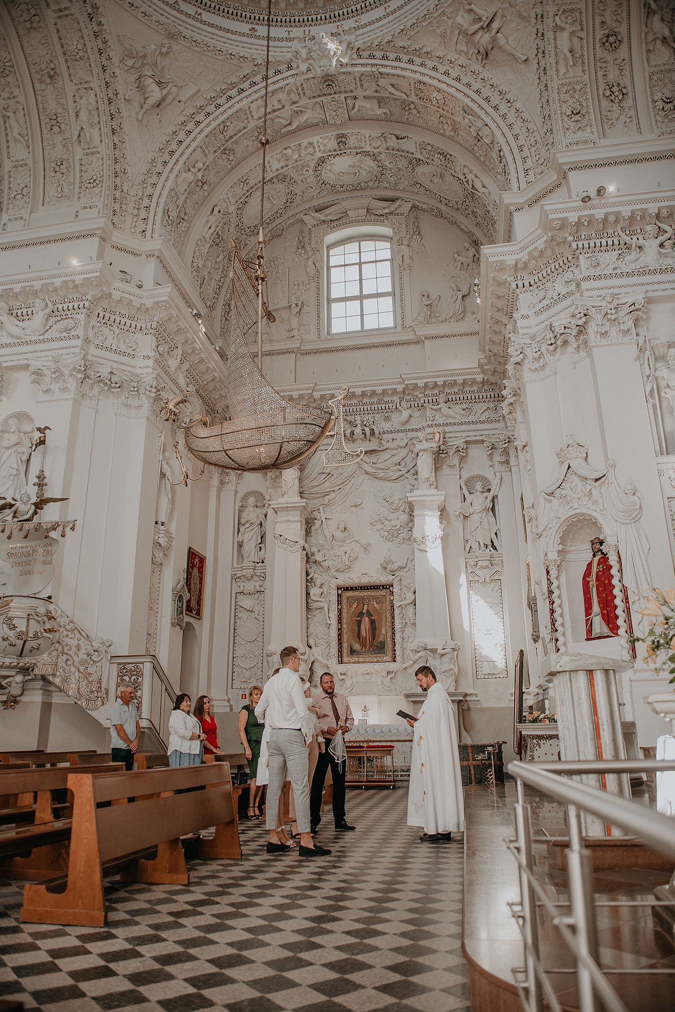 Interior panorama of an ornate baroque church with white walls, decorative plasterwork, and a black and white checkered floor.