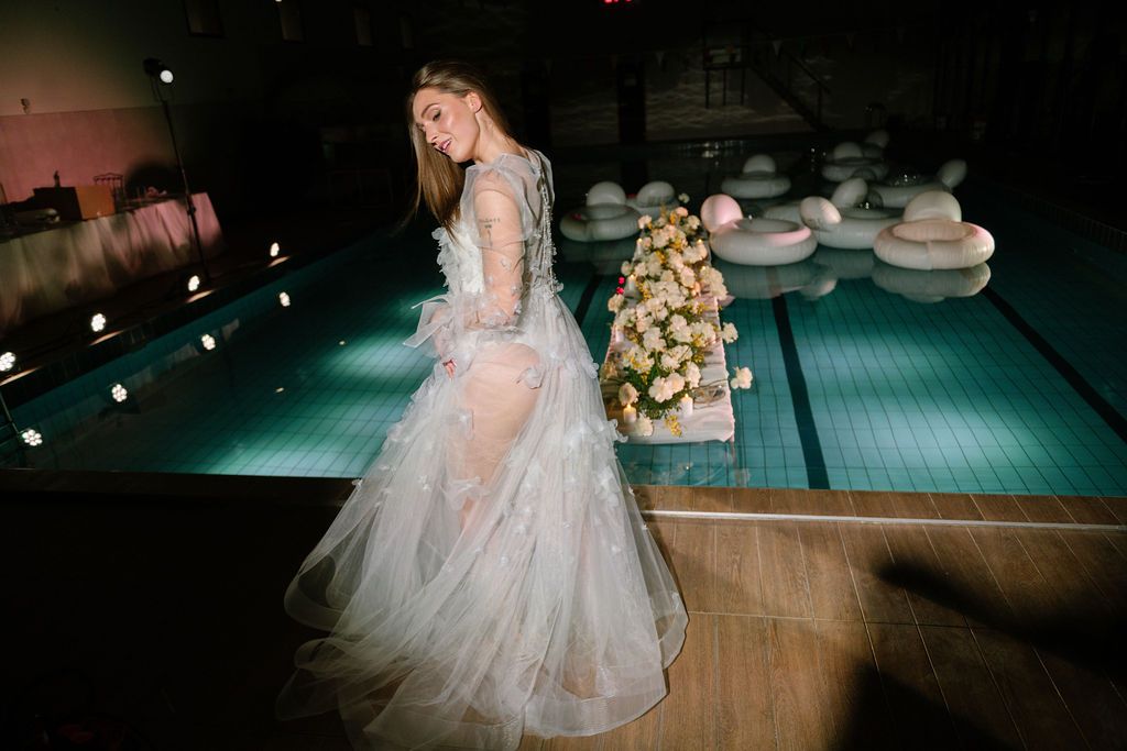 Bride in a sheer, flowing tulle wedding gown poses beside an indoor pool at night, with a floral arrangement and floating decor nearby.
