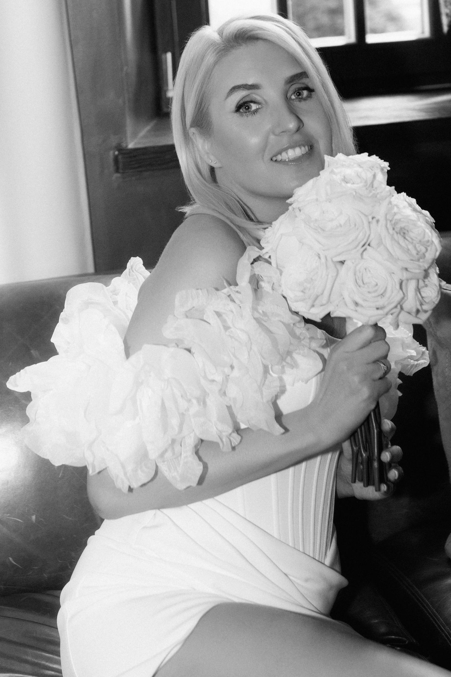 Black and white portrait of a smiling blonde bride in a white corset dress with ruffled sleeves holding a white rose bouquet.