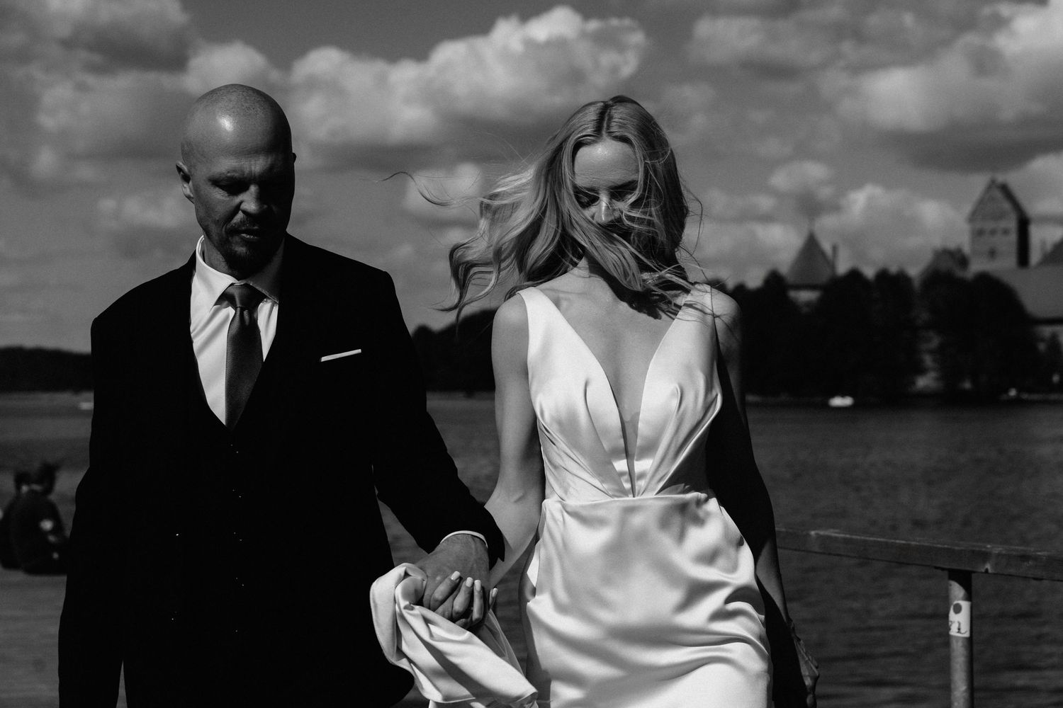 Black and white photo of a bride in a plunging satin gown walking hand in hand with a groom in a dark suit beside a lake.