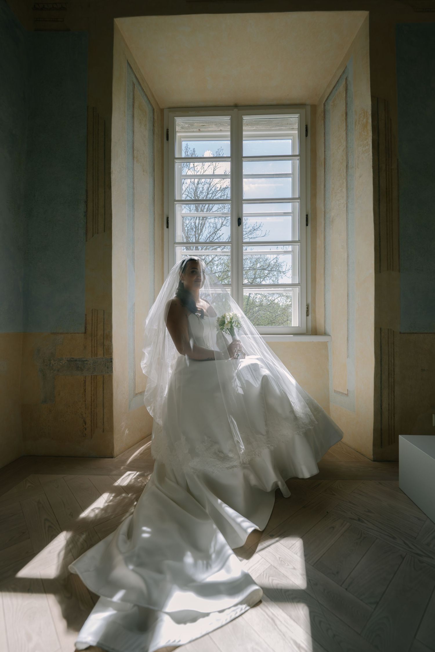 Bride in a flowing white gown and long veil sits by a tall window with sunlight streaming across the floor.