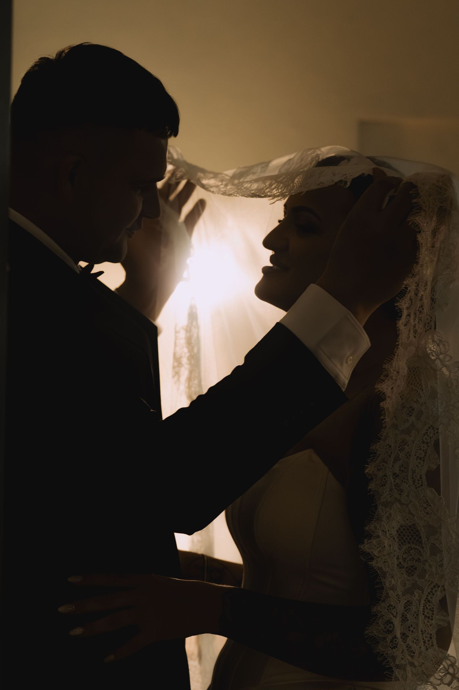 Silhouette of bride and groom sharing romantic moment, groom lifting lace veil with backlit golden glow behind them.