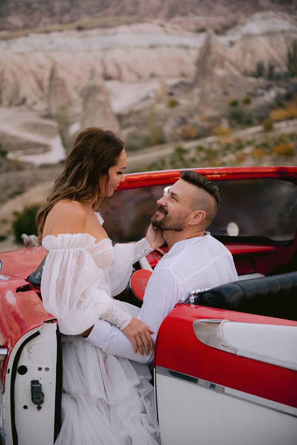 Couple sharing a romantic moment by a vintage red convertible in Cappadocia's scenic landscape.