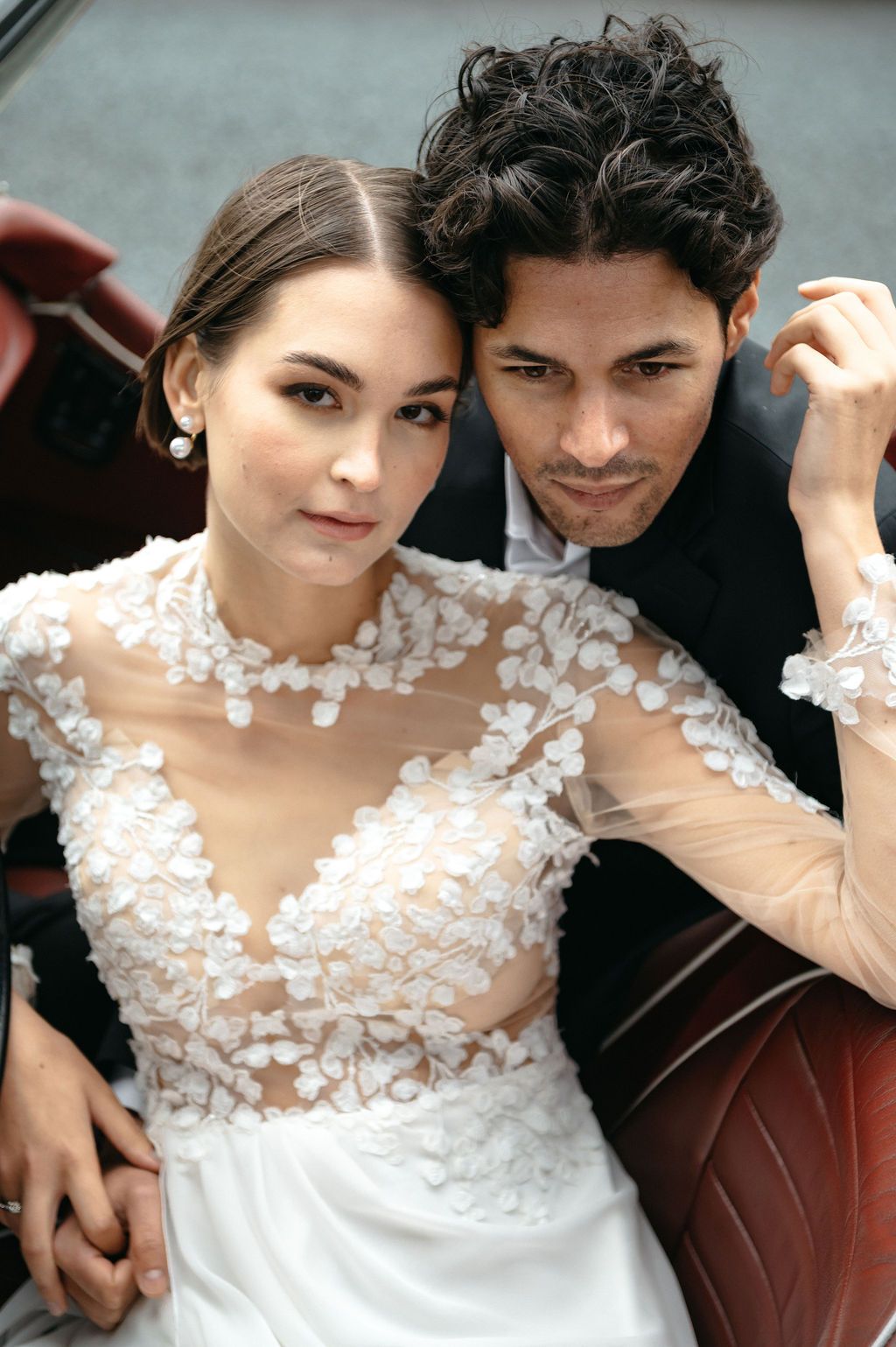 Bride in sheer lace wedding gown poses with groom in black suit inside a vintage car.