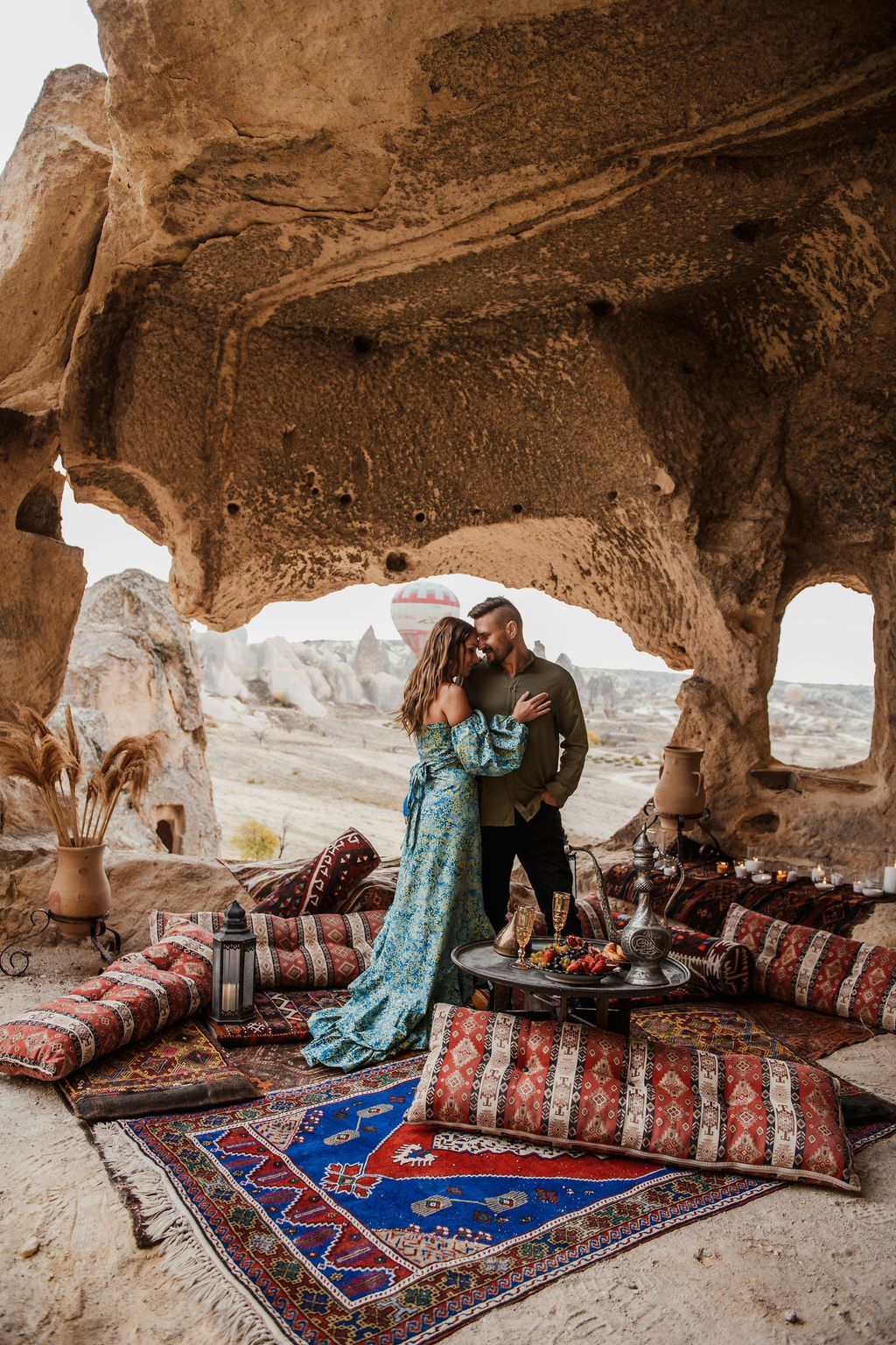 Couple embracing in a stunning Cappadocia cave setting with vibrant Turkish rugs and dramatic rock formations behind them.