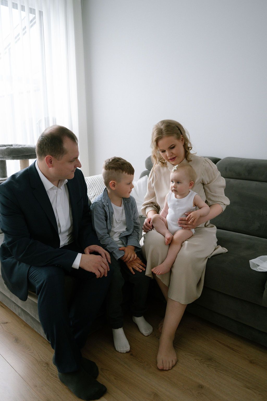 A family of four with two children sits together on a gray sofa in a bright living room.