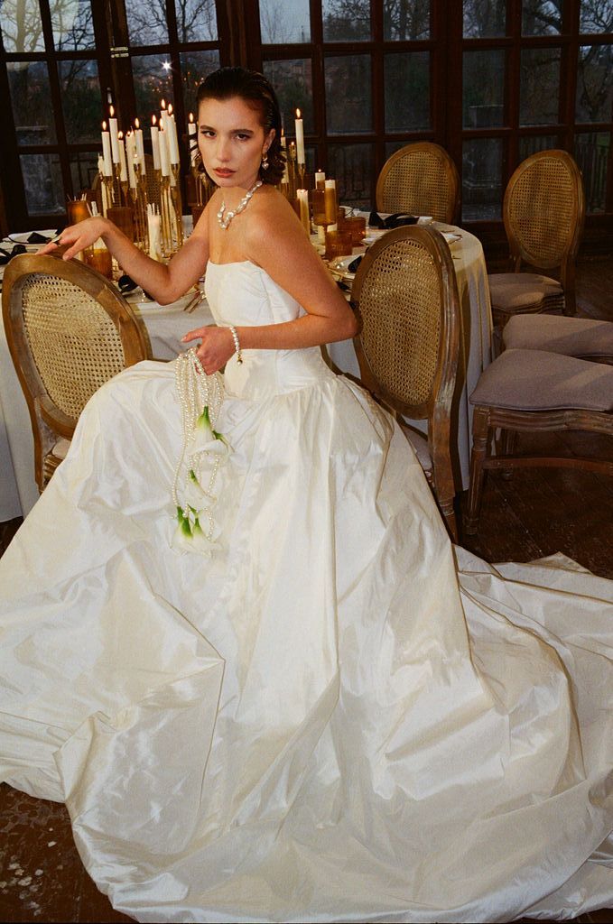 Bride in strapless ball gown holding white roses seated at candlelit table with pearl jewelry.