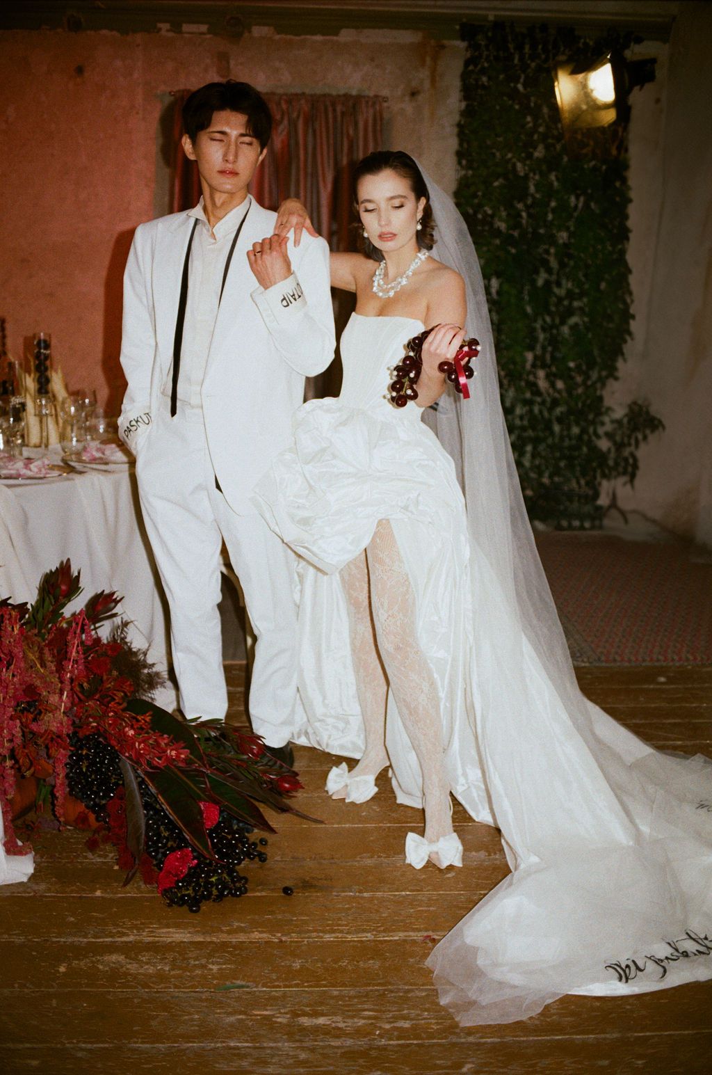 Elegant couple in white wedding attire pose at reception, bride in strapless gown with veil, groom in white suit.