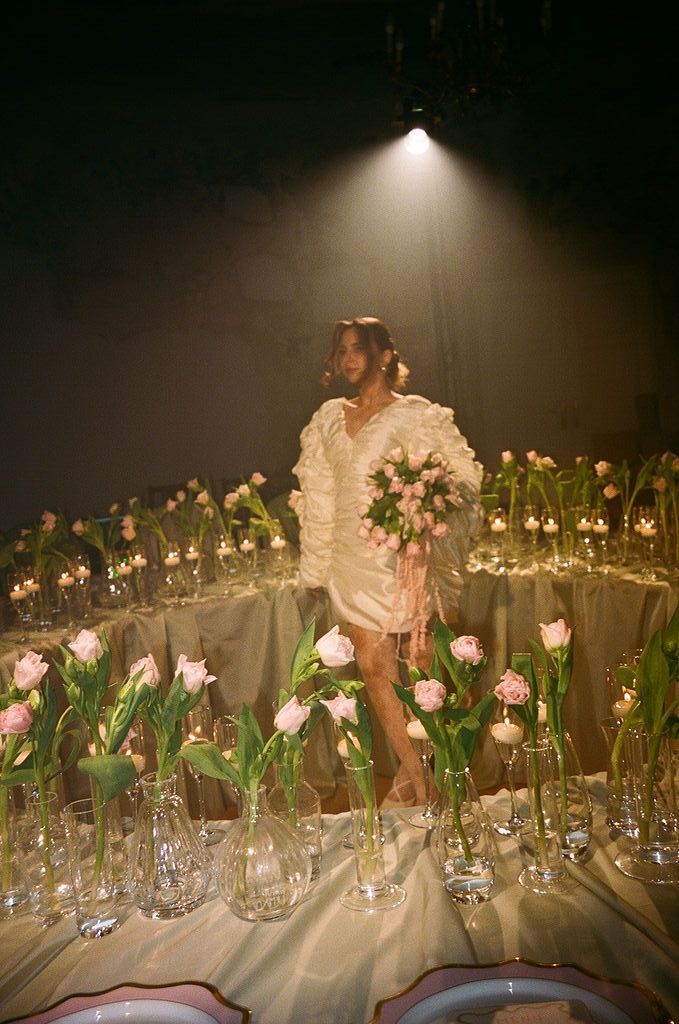 A woman in a white ruffled dress holds pink tulips surrounded by glass vases of tulips on a candlelit stage.