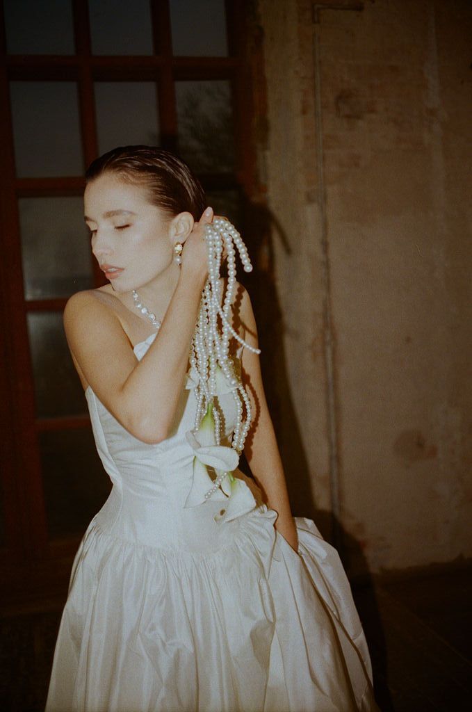 Bride in white ballgown with long pearl drop earrings poses gracefully with eyes closed in dimly lit venue.