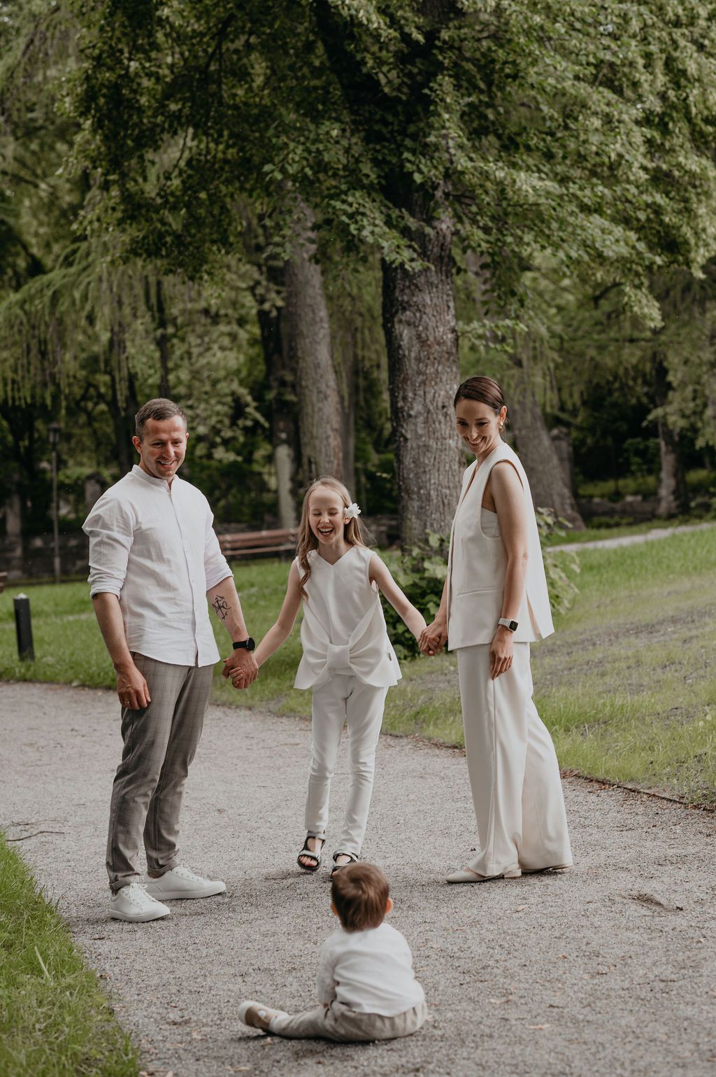 Family of four in white outfits enjoying a sunny day in a lush green park, with a toddler sitting on the path.