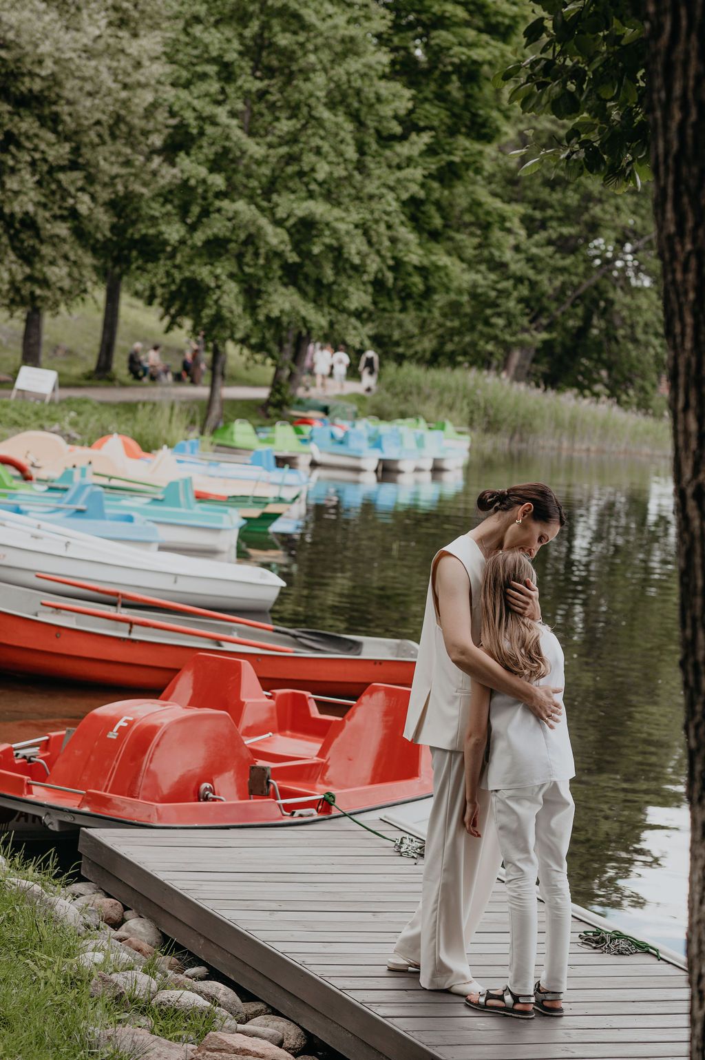 Couple embracing on a wooden dock beside colorful paddle boats on a calm lake in a lush green park.