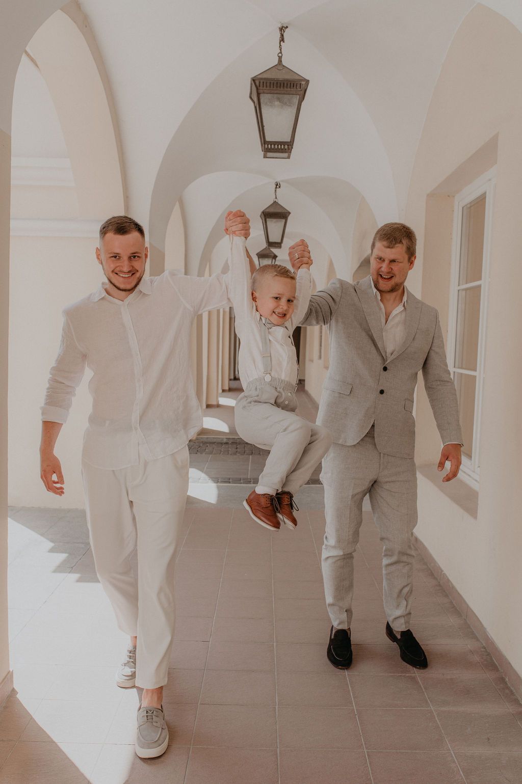 Two men in suits swing a laughing boy between them in a sunlit arched hallway.