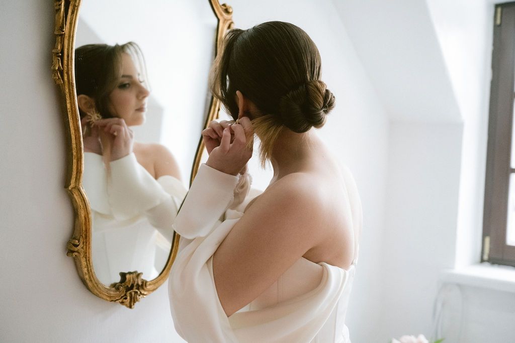 Bride in white gown putting on earrings before ornate gold mirror on wedding day.