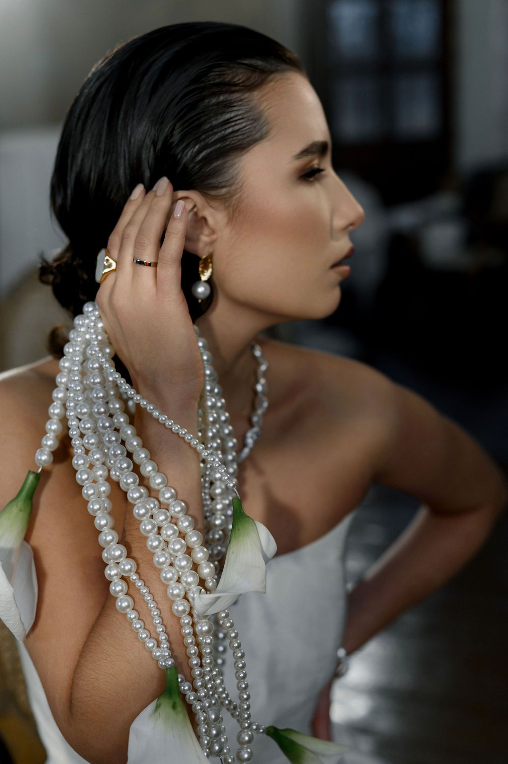 Woman in white dress wearing layered pearl necklaces and gold earrings, holding pearls near her face in elegant profile pose.