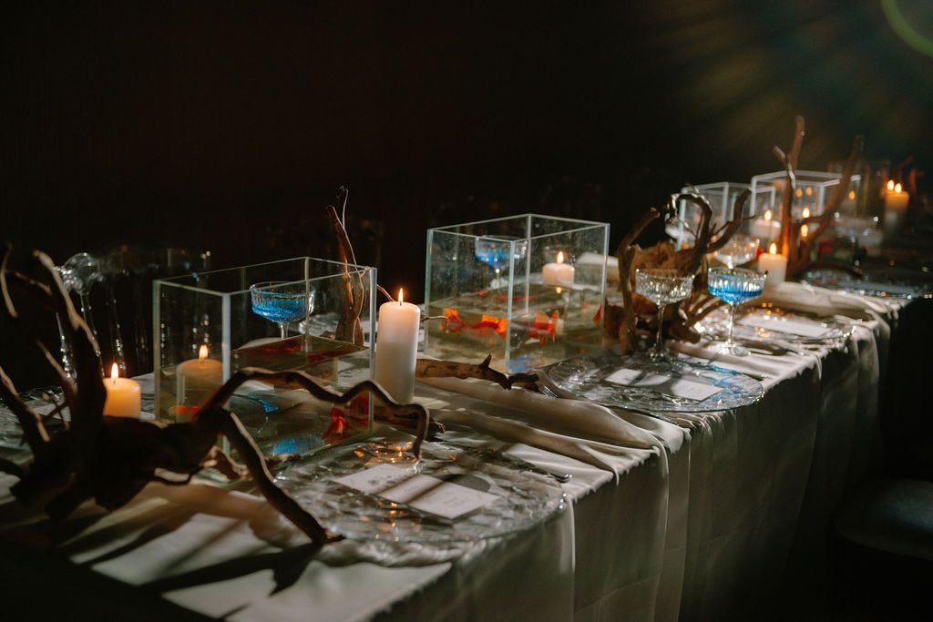 Elegant candlelit reception table with driftwood, glass terrariums, blue accents, and glowing candles for a moody event.