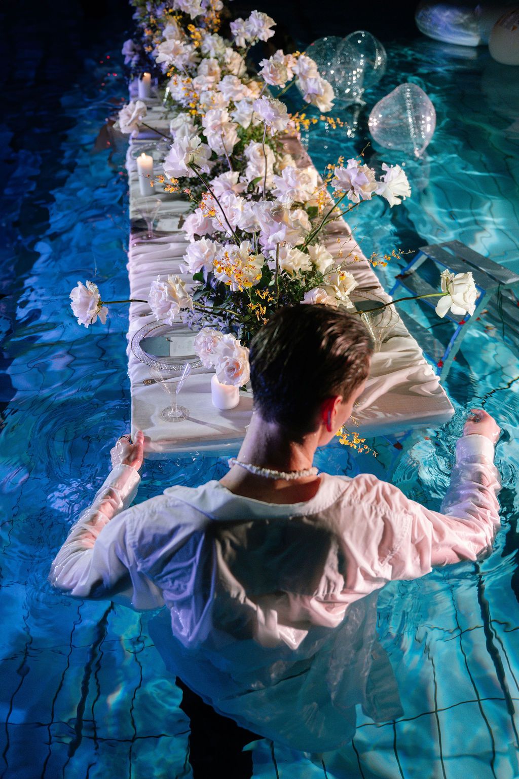Person in white shirt wearing pearl necklace stands in pool beside elegant floral table setting with white flowers.