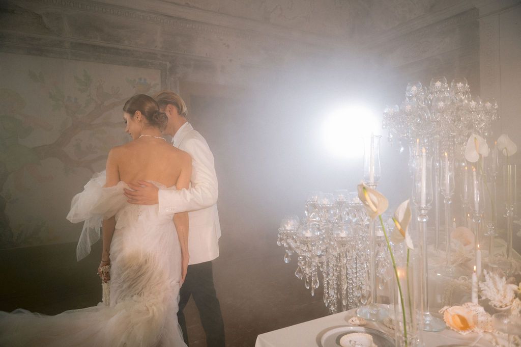Bride and groom share romantic dance in elegant chandelier-lit reception hall with misty atmosphere.