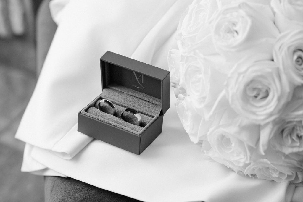 Black and white photo of wedding rings in a box beside a white rose bouquet.