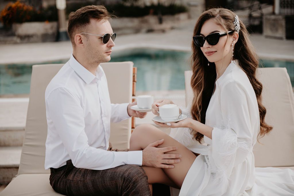 Elegant couple in white outfits sharing coffee by a pool, wearing stylish sunglasses.