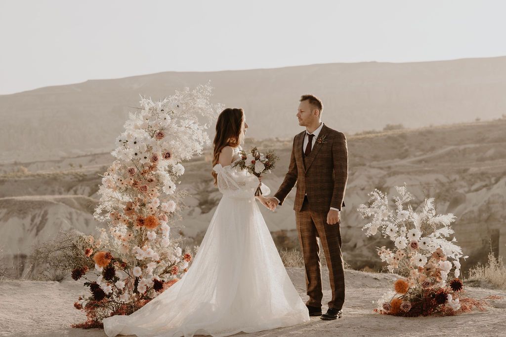 Bride and groom hold hands at floral arch with white and rust blooms in desert landscape.