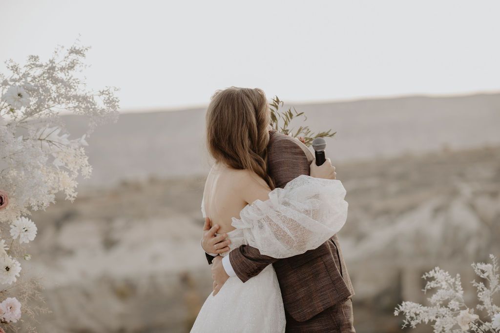 Bride and groom share a tender embrace near a white floral arch with a scenic desert landscape backdrop.