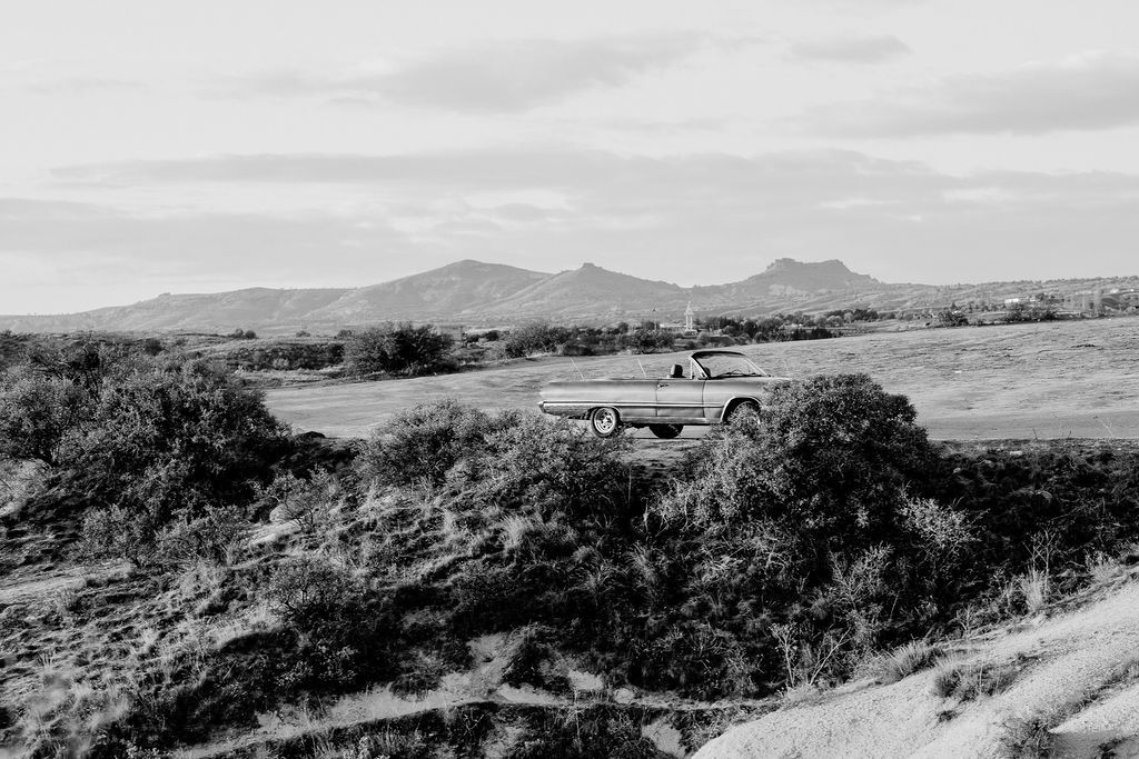 Black and white photo of a vintage truck driving through a rugged desert landscape with mountains in the background.