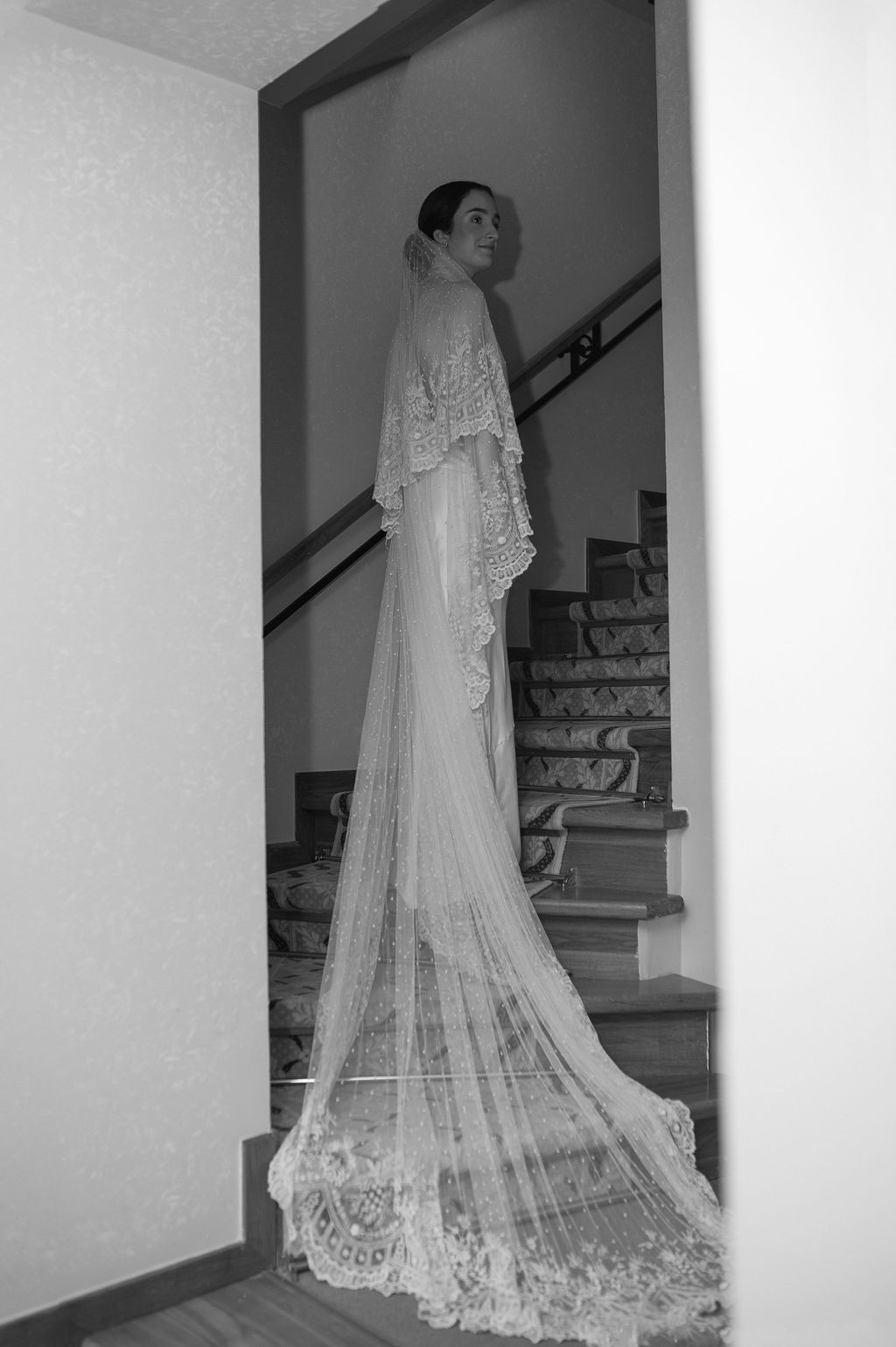 Bride in elegant lace wedding gown with long cathedral veil stands near staircase in black and white photo.