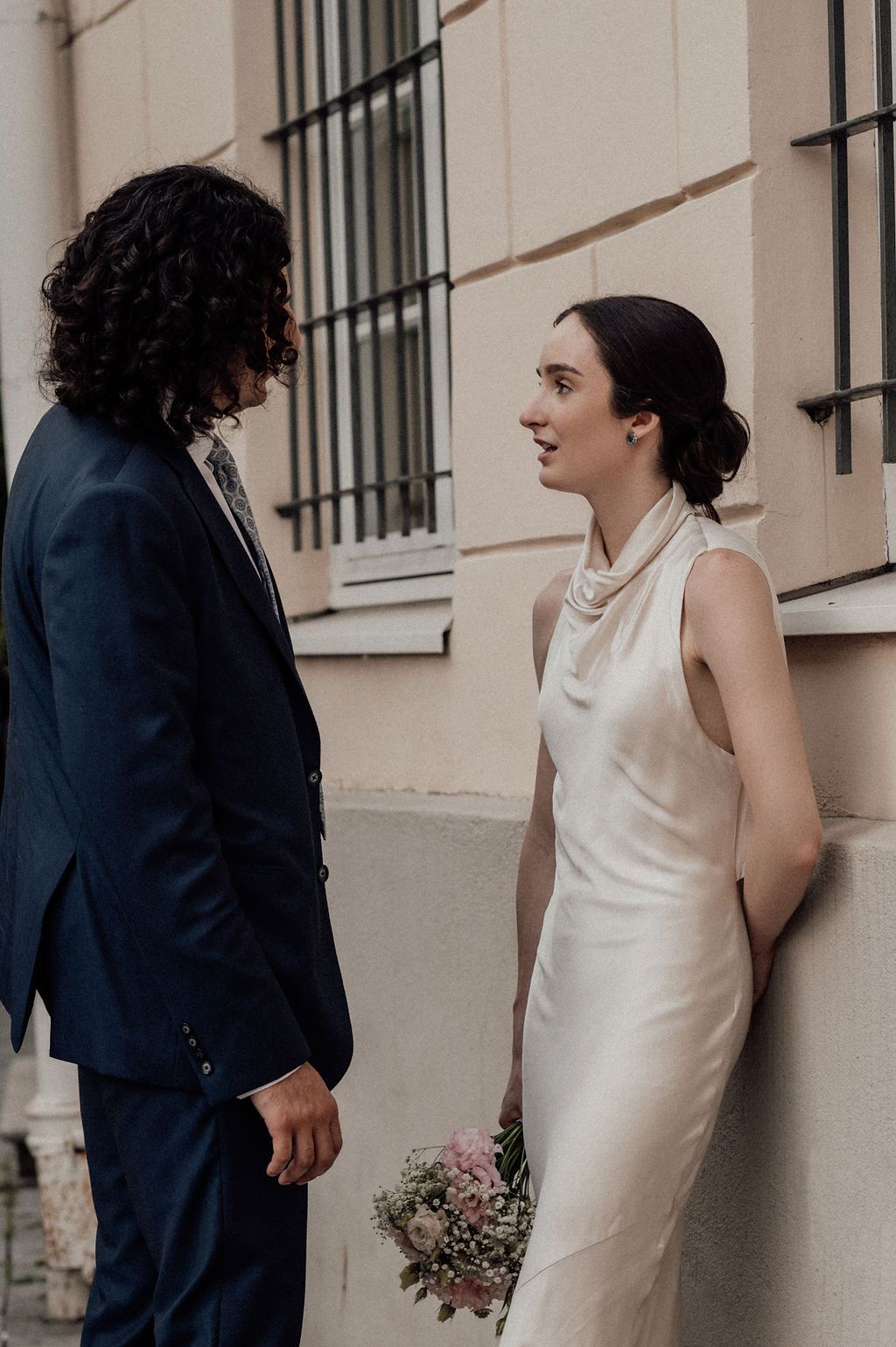 A bride in a white satin gown holds pink flowers while facing her groom in a navy suit against a beige wall.