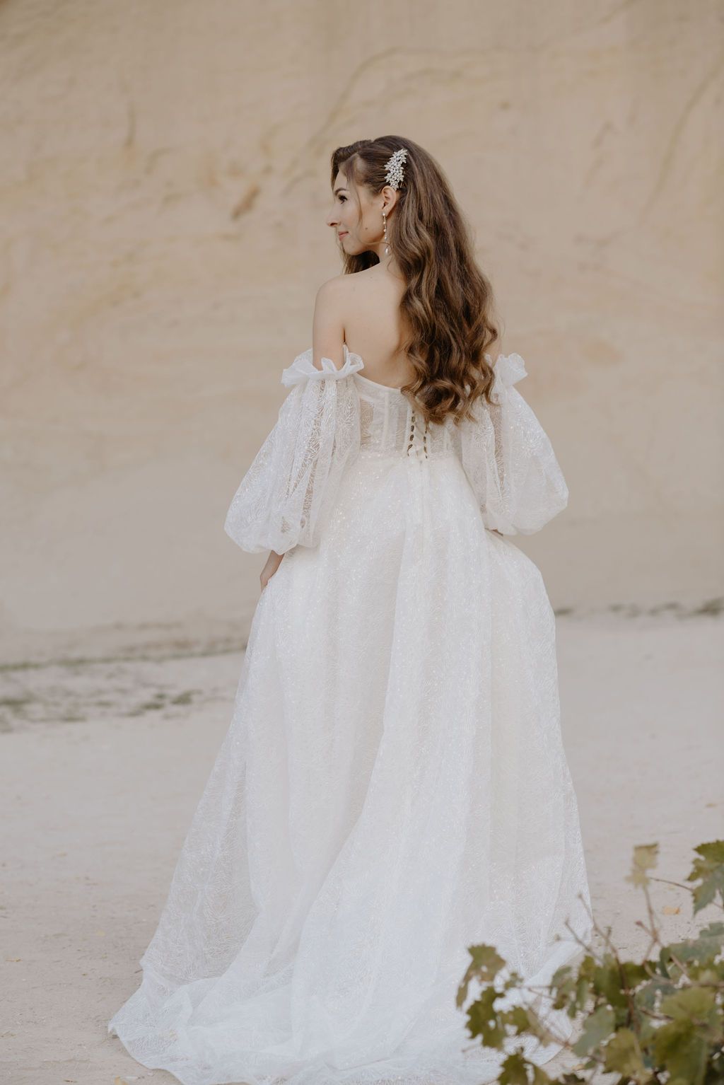 Bride in white off-shoulder lace wedding gown with puff sleeves and floral hair accessory, viewed from behind.