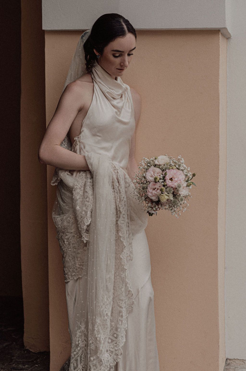 Bride in silk halter wedding gown with lace veil holds soft pink bouquet, posing elegantly against a blush wall.