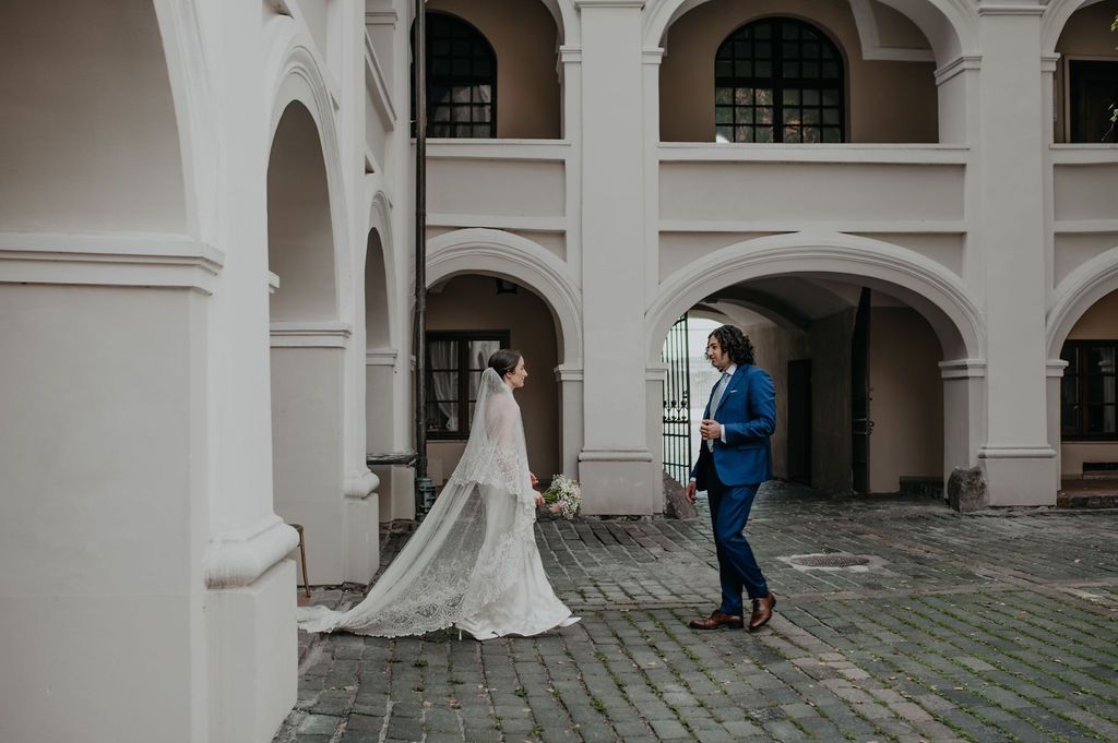 Bride in white gown and groom in blue suit share a romantic moment under arched colonnade during wedding photo session.