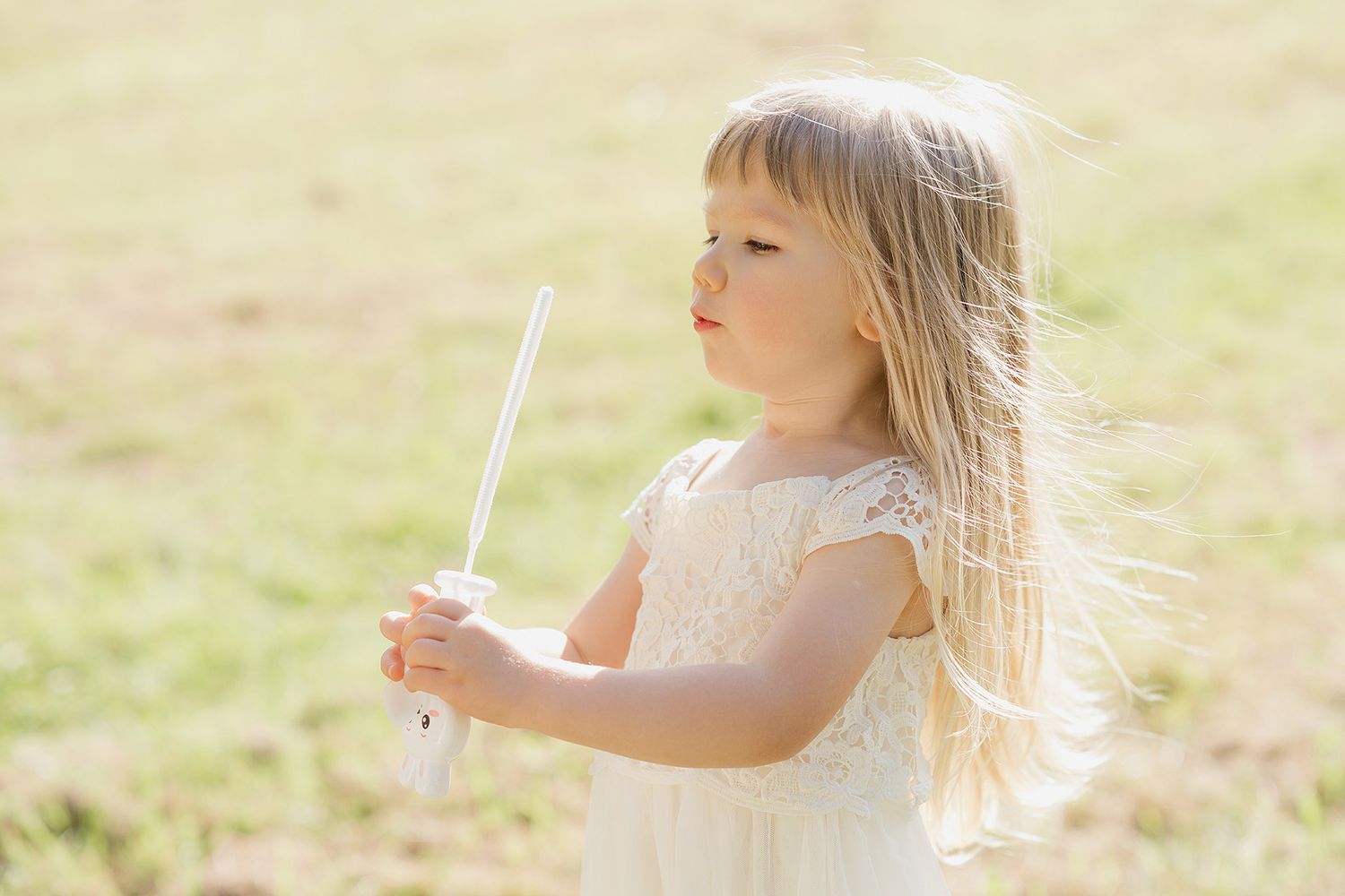 A child in a white dress blows dandelion seeds in a sunlit meadow, creating a magical moment in nature.