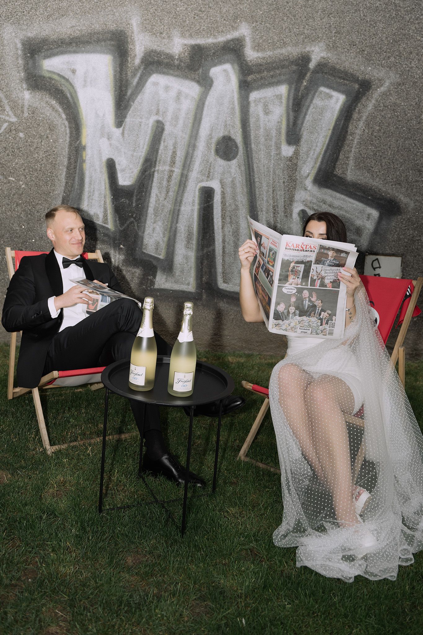 A wedding couple sits in lawn chairs against graffiti wall, reading newspapers with champagne bottles on a side table.