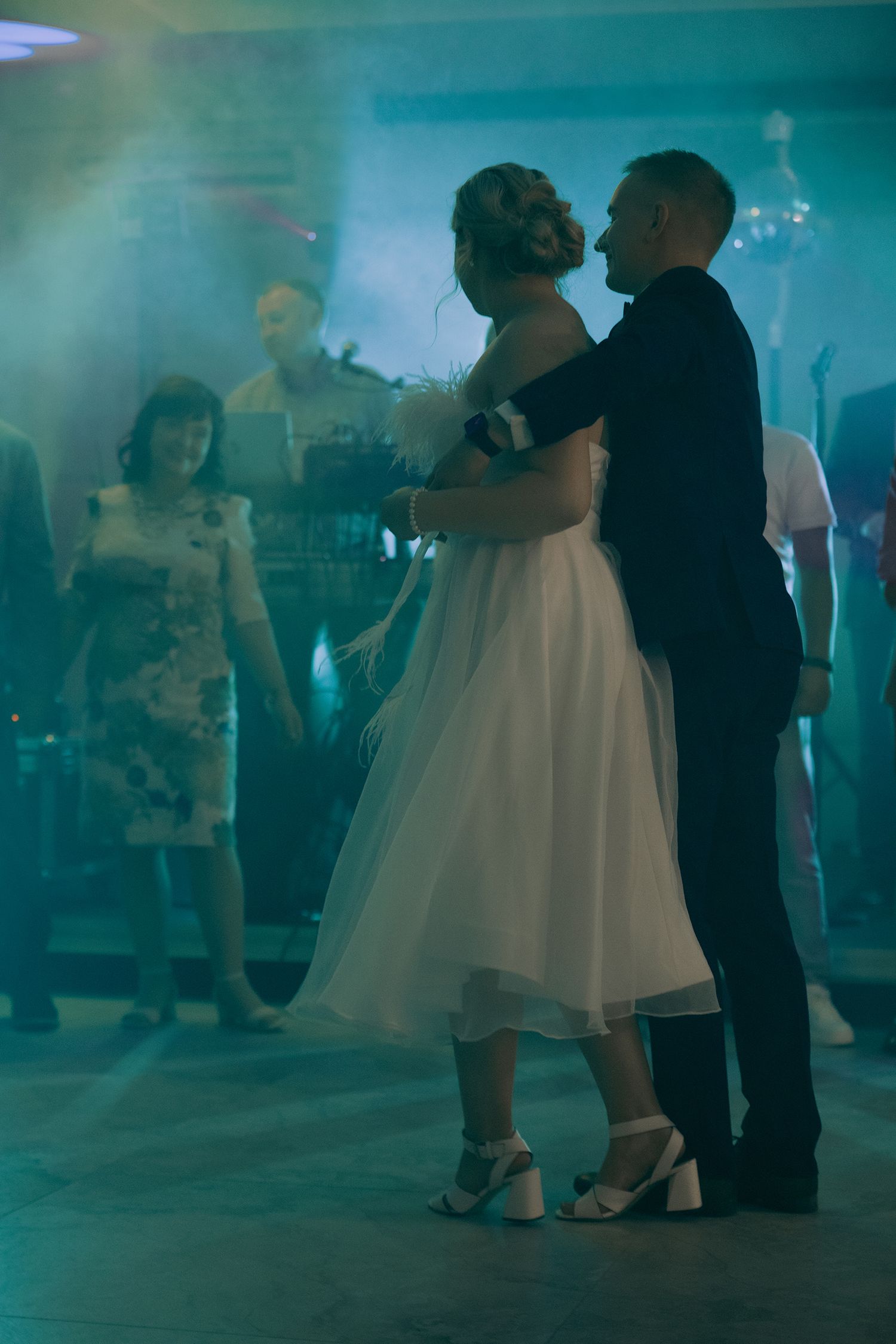 People dancing at a wedding reception in a dimly lit venue with blue-green lighting and hazy atmosphere.