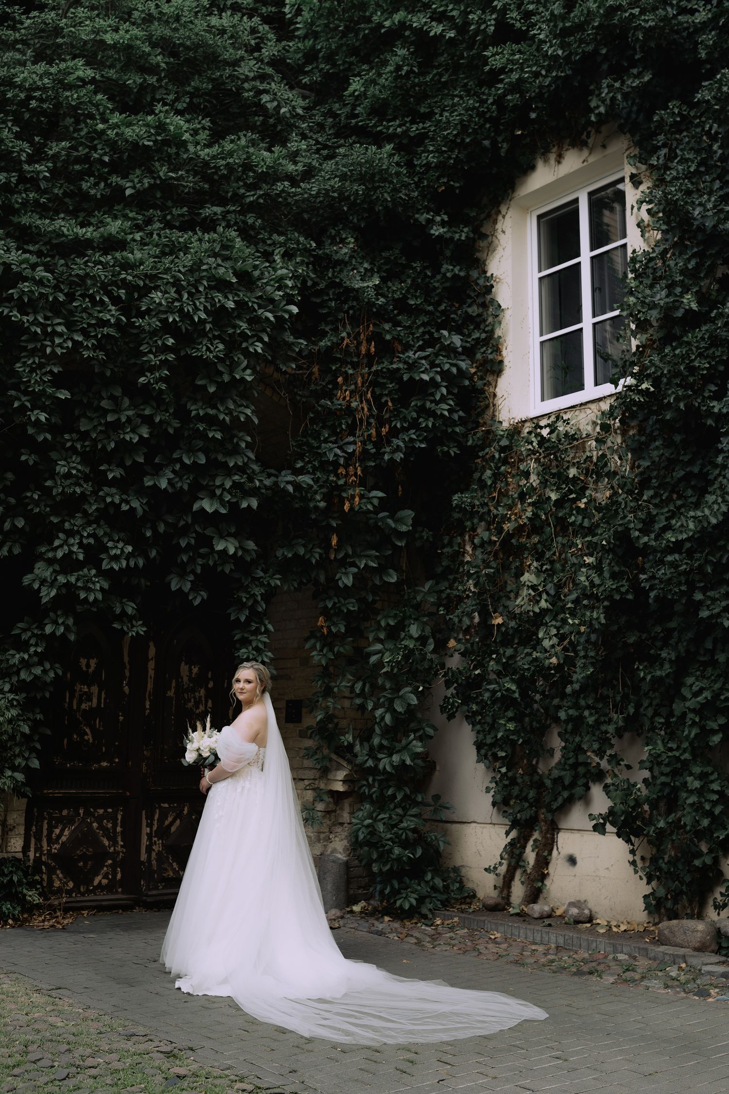 A bride in a flowing white wedding gown poses against an ivy-covered wall in a romantic garden setting.