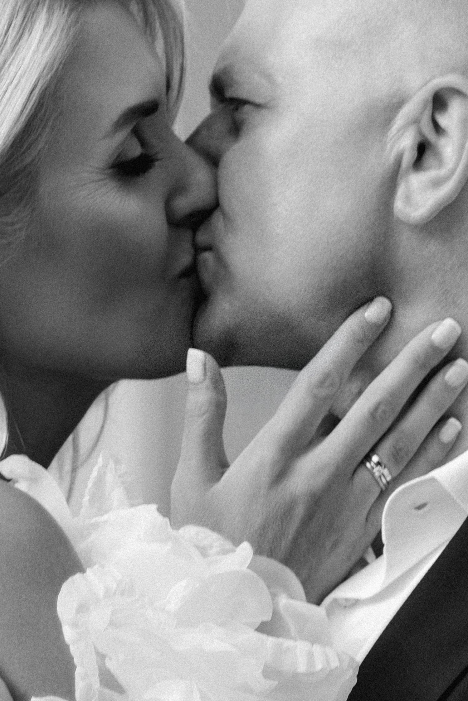 Black and white close-up of a bride and groom kissing, her hand with a wedding ring resting on his neck, a white floral bouquet below.