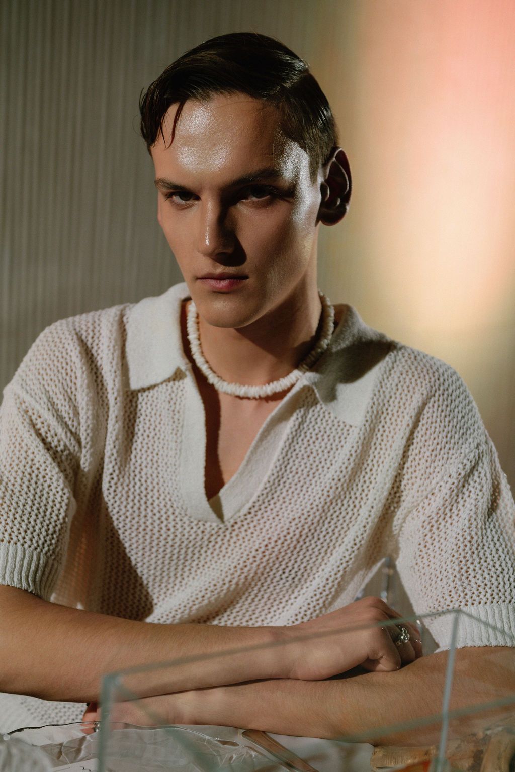 Young man with slicked-back dark hair wears a cream open-knit polo shirt and beaded necklace, posing with a serious look and arms crossed.