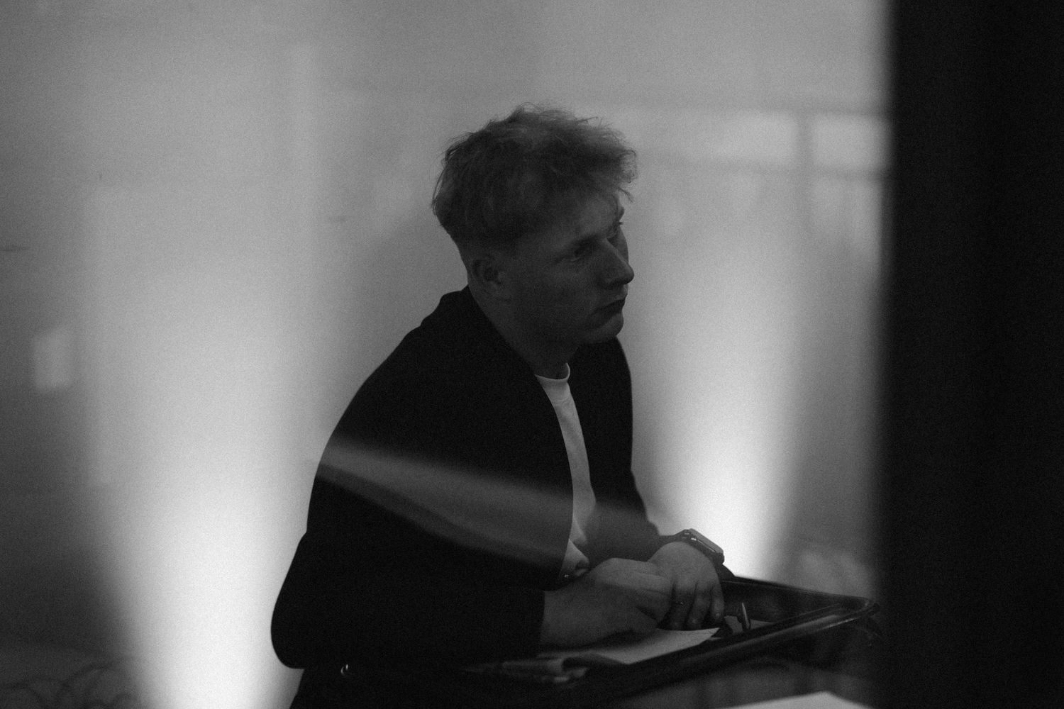 Black and white photo of a man in a dark jacket and white shirt seated at a table, looking to the side in soft, moody lighting.