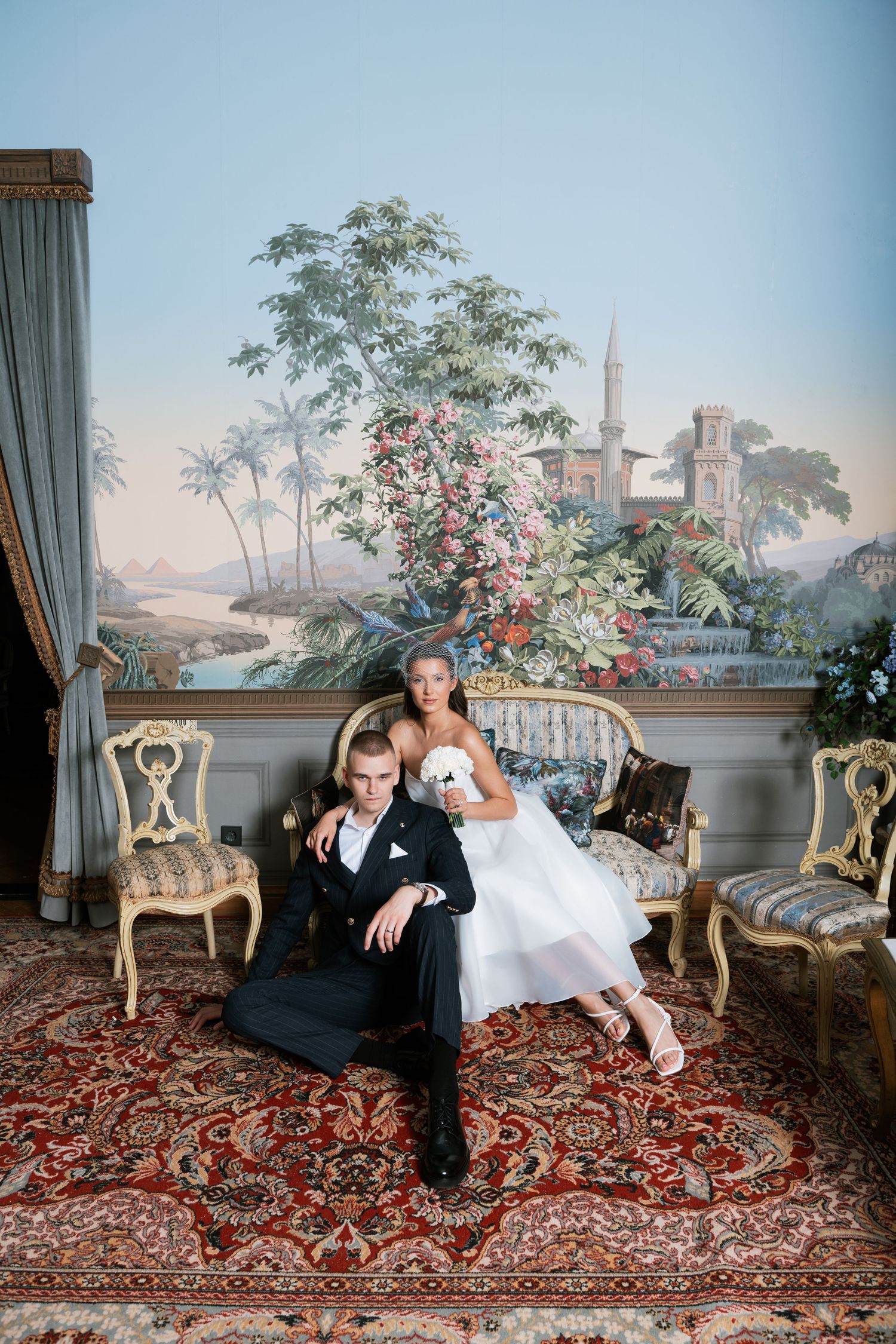 Bride in a white tulle gown holding a bouquet sits beside a groom in a dark suit on an ornate sofa before a tropical landscape mural.