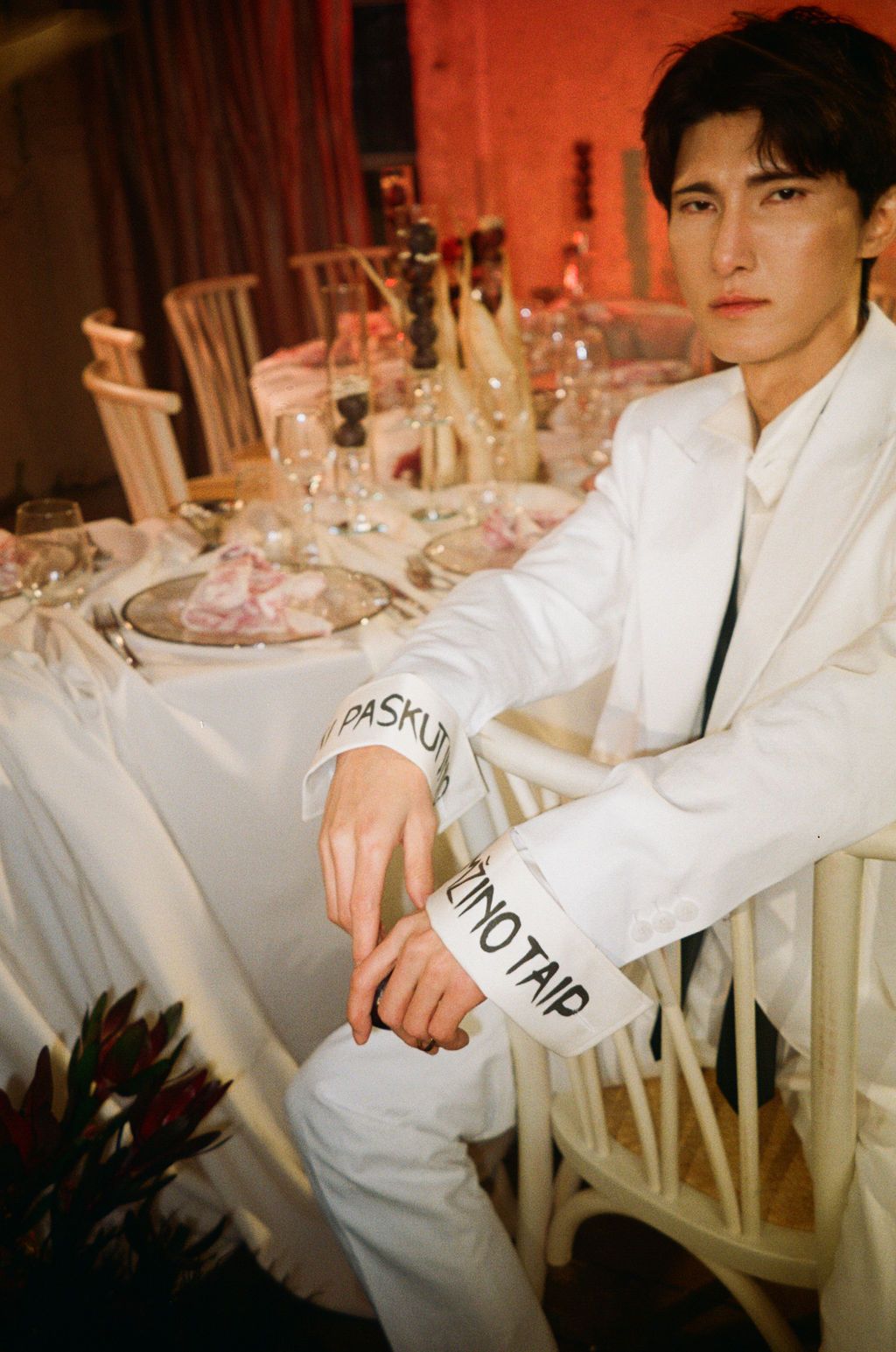 Man in white suit with branded cuffs sits at an elegantly set dining table with glassware and floral decor.
