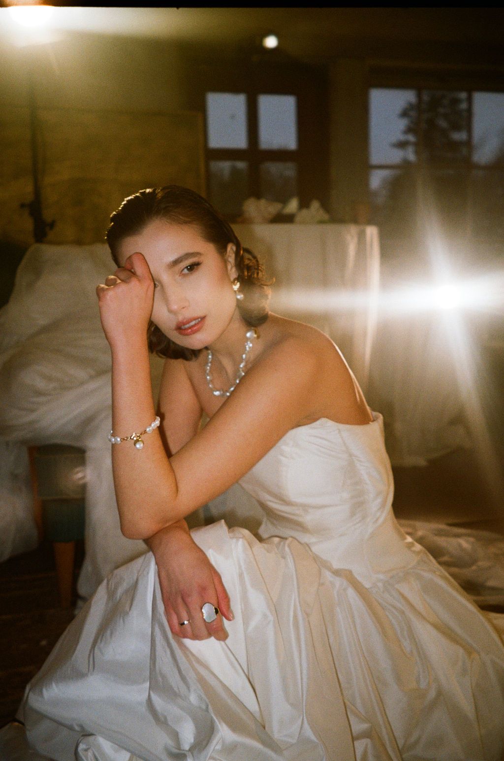 A bride in a white strapless gown sits gracefully, adorned with pearl jewelry, bathed in warm golden light.